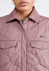 Prošívaná mauve bunda s límečkem, patentkami, dvěma náprsními kapsami a malým černým logem Nike na levé straně.