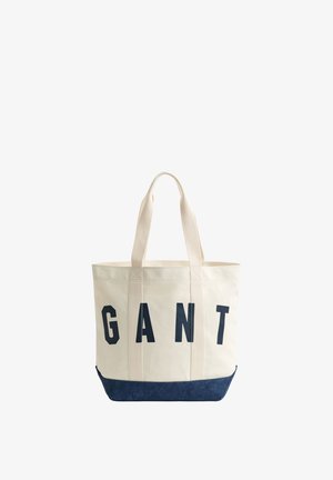 Beige Canvas-Tragetasche mit navyblauem Boden und großen schwarzen Buchstaben, die vorne "GANT" schreiben, ausgestattet mit zwei langen Schulterriemen.