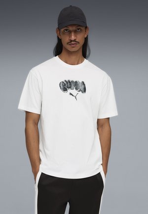 Uomo con capelli lunghi e baffi che indossa un berretto nero, una maglietta bianca con grafica e logo grigi, e pantaloni neri con strisce bianche, con le mani in tasca.