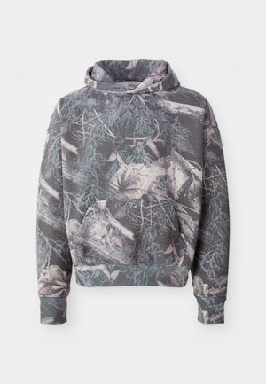 Haut à capuche gris avec un motif camouflage composé de branches et de feuilles dans des couleurs atténuées. Il possède une poche avant et un cordon de serrage sur la capuche.