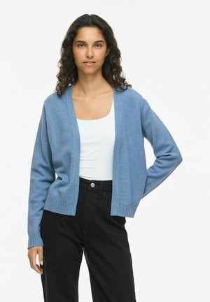 Vrouw met krullend donker haar, die een lichtblauwe open cardigan draagt over een wit topje en zwarte hoge taillebroeken, staand met één hand op de heup.
