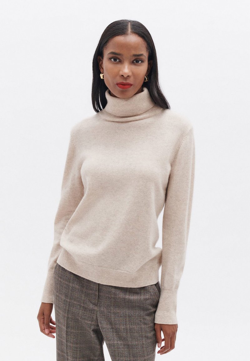 Beige Rollkragenpullover aus weichem Material mit langen Ärmeln, einer taillierten Passform und gerippten Akzenten an den Bündchen und dem Saum.