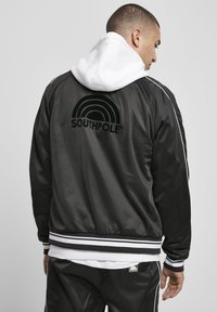 Giacca bomber nera con strisce bianche e logo. Presenta un collo alto, polsini elasticizzati e una trama liscia. Indossata sopra un hoodie bianco.
