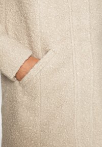 Beige tekstureret frakke med blødt, snoet stof, sidelommer og synlige sømme. Designet har en lige silhuet og ingen synlige lukninger.