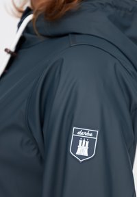 Marineblaue wasserdichte Jacke mit glatter Textur, Kordelzugkapuze und einem Schildpatch mit Schlossdesign sowie 'derbe'-Branding auf dem Ärmel.