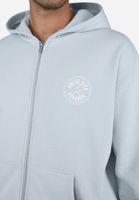 Lys blå hettegenser med glidelås og myk tekstur, frontlommer i kenguru-stil, og en sirkulær hvit logo på venstre brystområde.