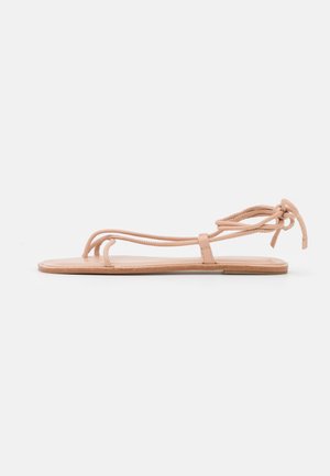 Flad beige sandal med tynde stropper og snørelukning ved anklen, minimalistisk design, lagt fladt på hvid baggrund.