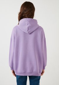 Sudadera lavanda de corte amplio con una gran capucha, puños y dobladillo acanalados. Tela suave con una textura lisa. La vista trasera muestra hombros caídos.