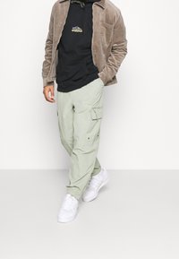 Giacca in corduroy tan sopra un hoodie nero, abbinata a pantaloni cargo verde chiaro e sneakers bianche. Presenta dettagli delle tasche e una vestibilità comoda.