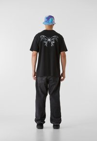 T-shirt noir avec un graphique de papillon blanc au dos. Associé à un jean sombre, ample et un chapeau seau tie-dye bleu.