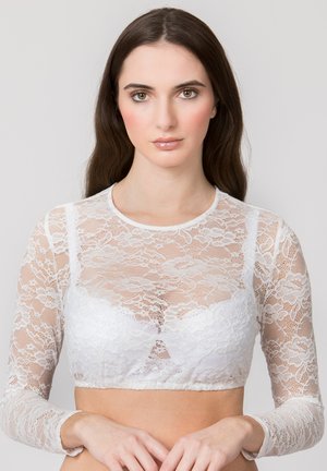 Frau mit langen braunen Haaren, die ein weißes langärmliges florales Spitzen-Crop-Top trägt, steht vor einem schlichten hellen Hintergrund.