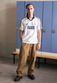 Maillot de sport blanc avec des accents bleus et jaunes, pantalon cargo beige et bottes noires, se tenant dans un vestiaire avec des casiers bleus.