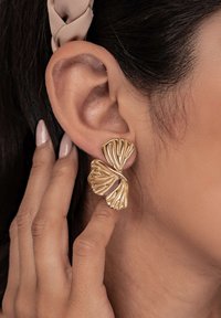 Primer plano de una mano tocando una oreja que lleva un pendiente dorado, texturizado, con forma abstracta y tres elementos parecidos a pétalos.