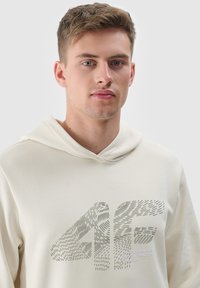 Beige hoodie met een getextureerd grafisch logo in groen en wit. Voorzien van een capuchon met trekkoord en geribbelde manchetten. Gladde stof, casual pasvorm.