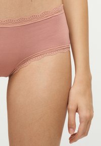 Vue latérale d'une personne portant des sous-vêtements roses bordés de dentelle, la main droite détendue le long de la cuisse, sur un fond uni.