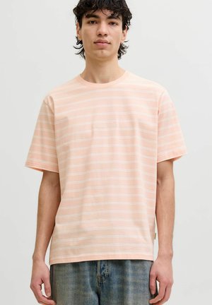 Ung mand med mørkt krøllet hår iført en lys ferskenfarvet stribet t-shirt og blå jeans mod en ensfarvet lys baggrund.