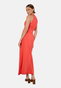 Robe maxi corail avec un bustier ajusté, une taille ceinturée et une jupe fluide. Le matériau semble lisse, complété par des sandales à talons épais.