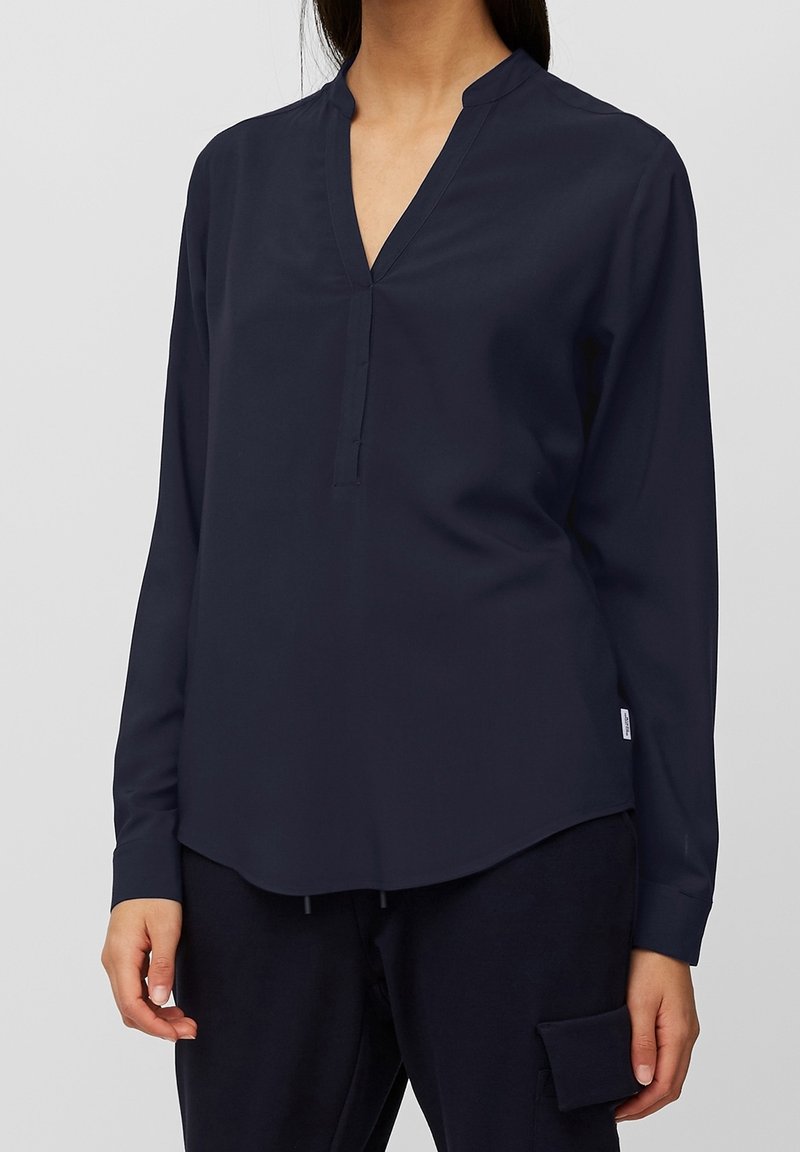 Blouse bleu marine à manches longues avec un col en V, une coupe ajustée et un ourlet arrondi. Confectionnée dans un tissu lisse à la texture brillante avec un minimum de garnitures.