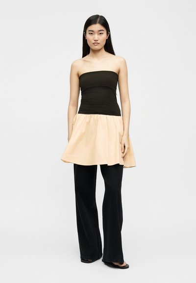 Γυναίκα με μακριά μαύρα μαλλιά, φοράει ένα ασύμμετρο μαύρο και μπεζ τοπ πάνω από μαύρο παντελόνι με φαρδιές παντελόνα, στέκεται μπροστά από απλό φόντο.