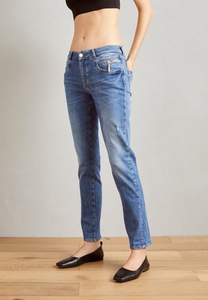 Schmal geschnittene blaue Jeans mit vorderer Reißverschlusstasche, getragen von einer stehenden Person in schwarzen Ballerinas auf einem Holzboden vor einer schlichten Wand.