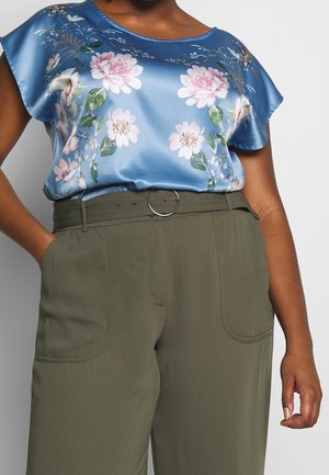 Femme portant un chemisier en satin bleu à motifs floraux, rentré dans un pantalon taille haute vert olive avec une ceinture assortie et une boucle ronde.