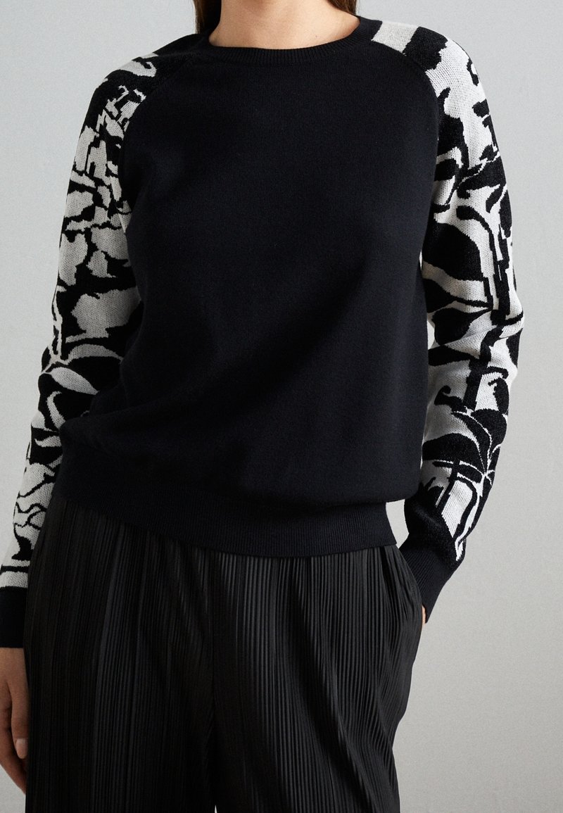 Femme portant un pull noir avec des manches à motifs abstraits noir et blanc et un pantalon plissé noir, debout devant un fond gris.