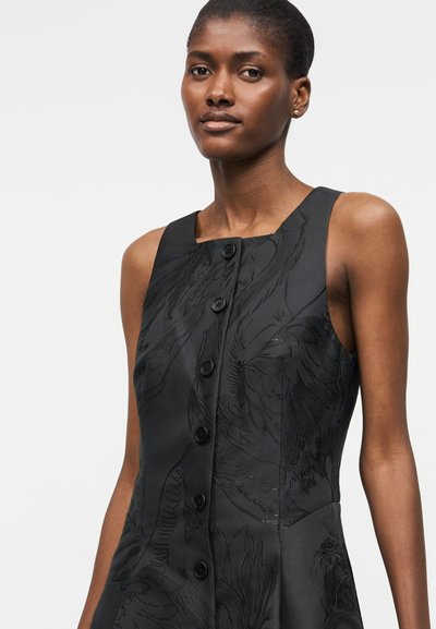 Γυναίκα που φοράει μαύρο αμάνικο φόρεμα με μεγάλα κουμπιά και διακριτικό floral σχέδιο, στέκεται μπροστά σε απλό φωτεινό φόντο.