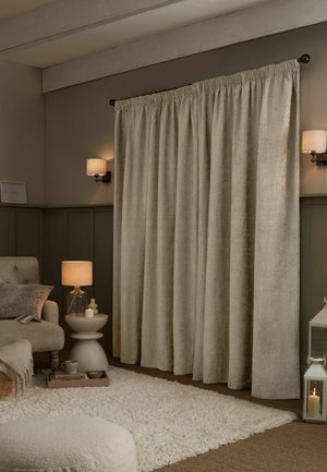 Tende beige chiuse appese a un’asta in un soggiorno accogliente con una lampada accesa, applique da parete, un tappeto morbido e una poltrona imbottita.