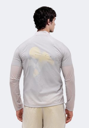 Άνδρας με σκούρα μπούκλα φοράει ανοιχτό γκρι μακρυμάνικο πουκάμισο με οπές αερισμού και μπεζ σορτς, με την πλάτη προς ένα απλό φόντο.