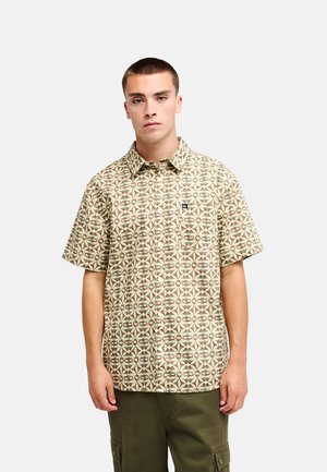 Młody mężczyzna w beżowej koszuli na krótki rękaw z geometrycznym wzorem i oliwkowo-zielonych spodniach typu cargo, stojący na białym tle.