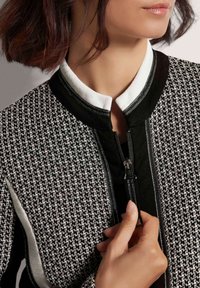 Reißverschlussjacke mit einem schwarz-weißen Hahnentrittmuster, strukturiertem Stoff, schwarzen Akzenten und einem weißen Kragen.