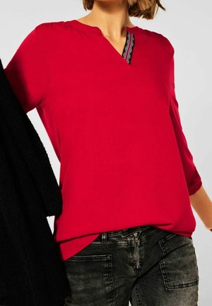 Person iført en lys rød top med lange ærmer og sort-hvide striber ved halsudskæringen, kombineret med mørkegrå jeans og holder et sort stykke tøj.