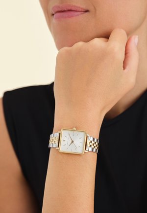 Personne portant une montre-bracelet à cadran carré bicolore or et argent avec un cadran blanc et des marquages minimaux au poignet gauche.