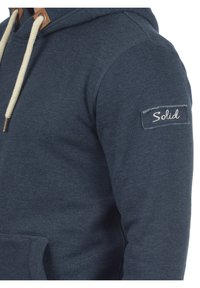 Marinblå hoodie i mjukt tyg, med en känguruficka, justerbar huva med beige snöre och en texturerad etikett med "Solid."