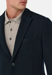 Tmavě modrý texturovaný blazer s výstřihem do klopy a jedním knoflíkem, kombinovaný se světle béžovým polokošilem se třemi knoflíky a jemnou pletenou strukturou.