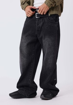 Personne portant un jean noir ample, des baskets noires et une veste verte, en train d'attacher une ceinture noire.
