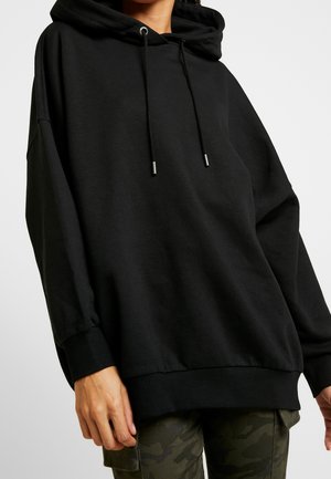 Osoba ubrana w luźną czarną bluzę z kapturem i sznurkami oraz spodnie w moro, stojąca na jednolitym jasnym tle.