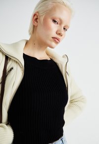 Juodas ribotas megztinis, vilkimas po kremu zip-up striuke. Audinys atrodo minkštas, matomi siūlai ir lygus tekstūra. Džinsiniai šortai dalinai matomi.