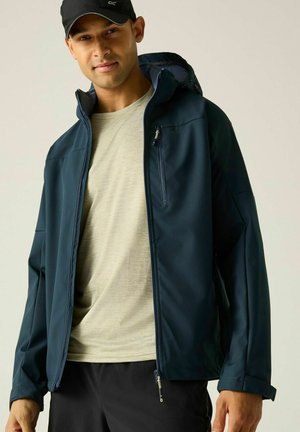 Hombre con gorra negra, chaqueta con cremallera azul marino, camisa beige y pantalones negros, de pie frente a un fondo claro y liso.