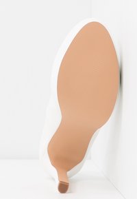 Chaussure blanche à talons hauts, inclinée, avec une semelle lisse marron clair et un talon étroit, appuyée contre une surface blanche aux coins.