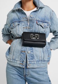 Czarna torba na pasek o fakturze z teksturą z metalowymi elementami w kolorze srebrnym, z zapięciem na klamrę, noszona na jasnoniebieskiej kurtce dżinsowej.