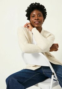 Elfenbeinfarbener Strickcardigan mit einem strukturierten Muster, langen Ärmeln und einem lässigen Schnitt, kombiniert mit dunkelblauen Jeans, sitzt auf einem weißen Stuhl.