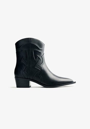 Botín de cuero negro hasta el tobillo con punta afilada, diseño texturizado en zigzag, tacón bajo y acabado suave. Estilo y detalles minimalistas.