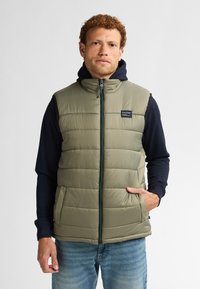 Padded olivgrüner Weste mit hohem Kragen, Frontreißverschluss und seitlichen Taschen. Getragen mit einem navyblauen Langarmhemd darunter.