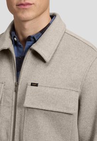 Homme portant une veste en laine beige avec fermeture éclair à l'avant et poche à rabat sur la poitrine, par-dessus une chemise à carreaux bleue et noire.