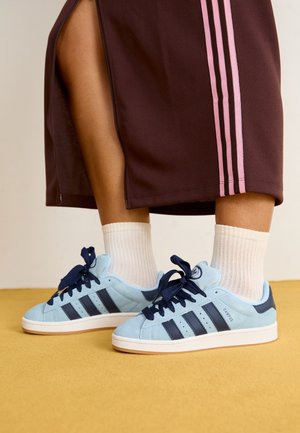 Personne portant des chaussettes blanches, des baskets Adidas Campus bleu clair avec des bandes bleu foncé et une jupe marron avec des bandes roses.