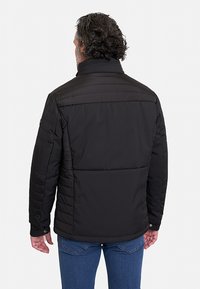 Chaqueta negra acolchada con cuello alto, sisa y detalles acolchados verticales. La tela parece suave y ligera.