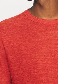 Pull rouge en tissu maille texturé. Présente un col ras du cou et un col côtelé. La surface affiche un motif subtil et varié.