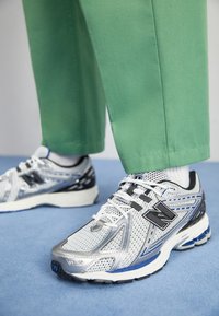 Person som bär vita och blå New Balance-sneakers med vita strumpor och gröna byxor, som står på ett ljusblått golv.