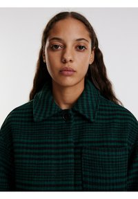 Groene en zwarte geruite wollen jas met een opvallende kraag, enkele knoopsluiting en een borstzak, met een gestructureerd patroon.